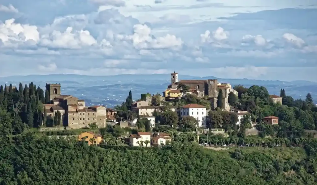 consulente di viaggio Panorama di un idilliaco paese collinare in Toscana, circondato da vegetazione lussureggiante e un cielo nuvoloso.