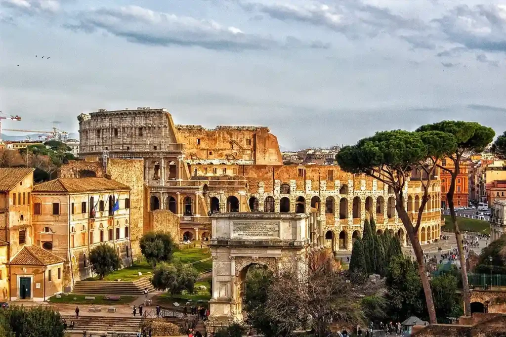 consulente di viaggio Vista del Colosseo e dell'Arco di Costantino a Roma, con pini marittimi in primo piano sotto un cielo nuvoloso.